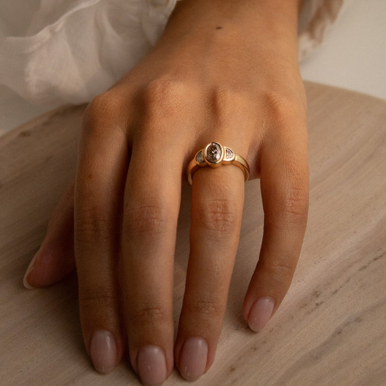 A gold ring with a round clear gemstone worn on a person's ring finger, shown with a white ruffled sleeve and a light wood surface in the background.