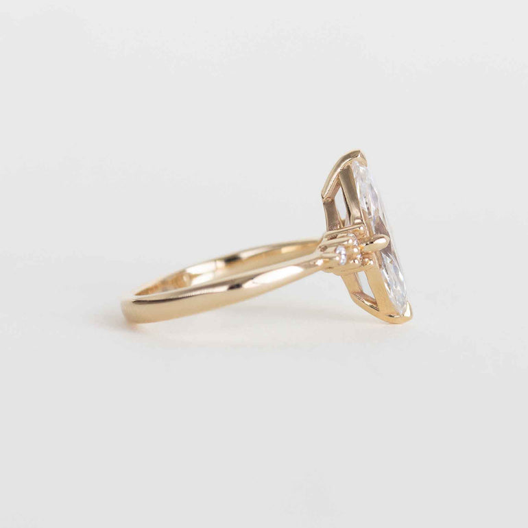 A gold ring featuring a large marquise-cut clear gemstone in a prong setting, with a small round accent stone on the band, photographed on a clean light gray background.