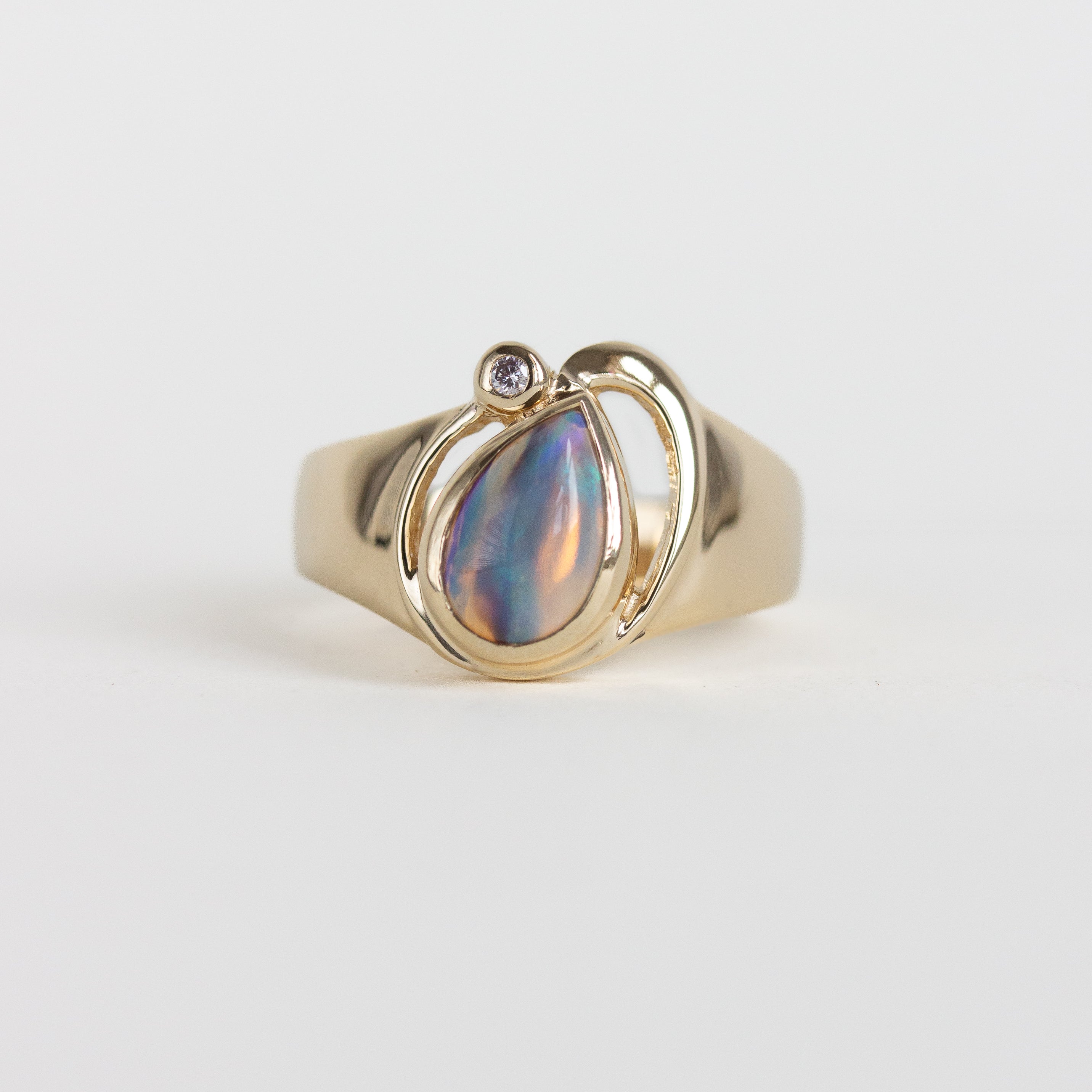 A gold ring featuring a teardrop-shaped iridescent shell inlay with a small round clear gemstone at the top, set against a plain light gray background.