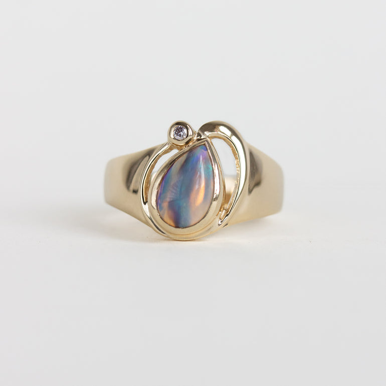 A gold ring featuring a teardrop-shaped iridescent shell inlay with a small round clear gemstone at the top, set against a plain light gray background.