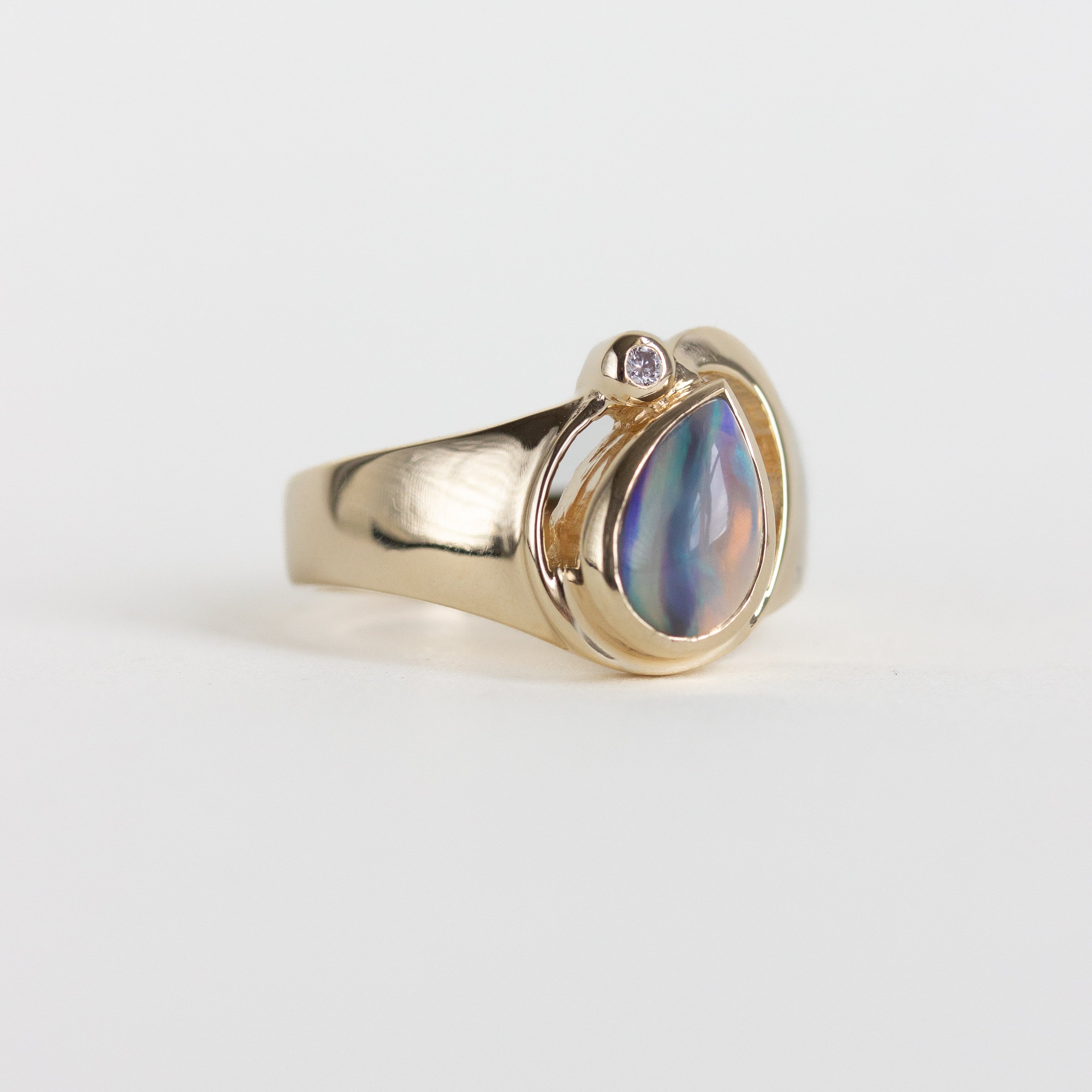 A gold ring featuring a teardrop-shaped iridescent shell bead with blue, purple, and green hues, accented by a small round clear stone on the side, set against a plain light gray background.