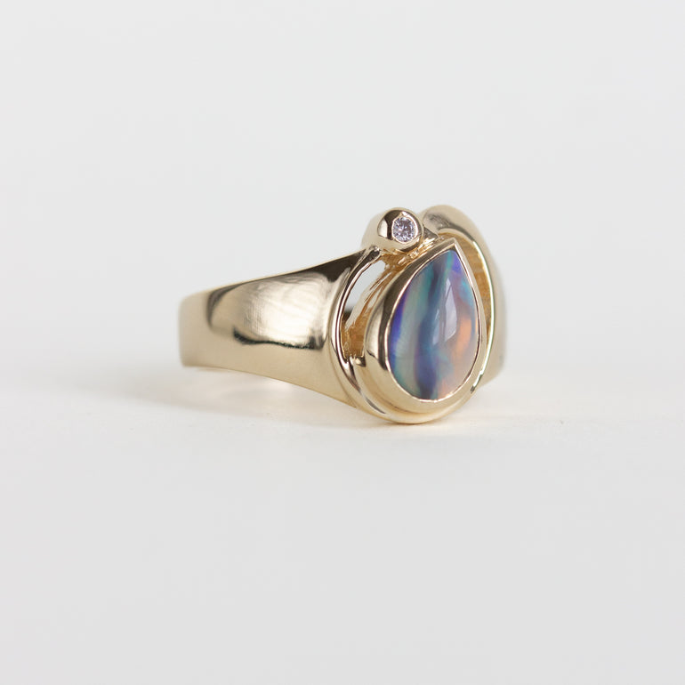 A gold ring featuring a teardrop-shaped iridescent shell bead with blue, purple, and green hues, accented by a small round clear stone on the side, set against a plain light gray background.