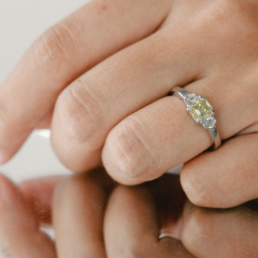 A close-up of a hand wearing a silver ring with a Asscher-cut yellow diamond, resting on a reflective surface.